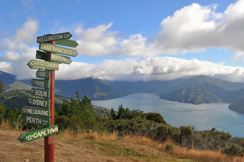 Directional signpost with global city distances overlooking a large body of water and mountains